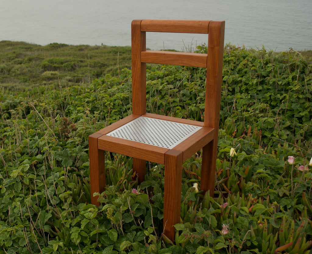 Modern minimalist designer wooden chair with a perforated metal seat. Front view with dense foliage and the ocean in the background.