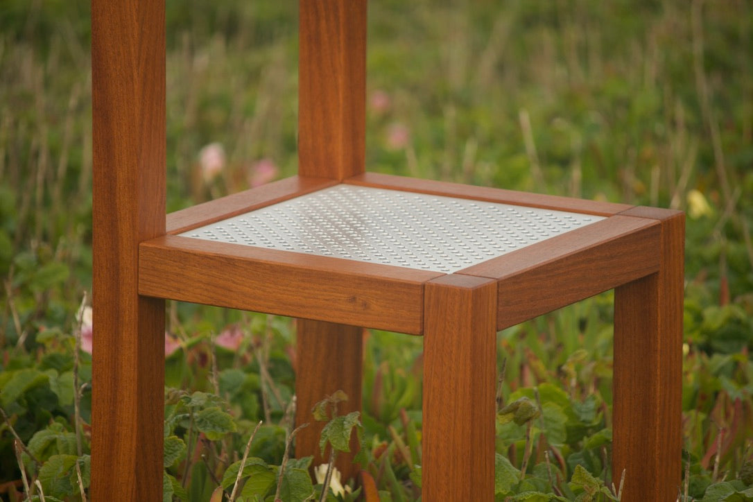 Modern minimalist designer wooden chair with a perforated metal seat. Front close-up view with dense foliage in the background.