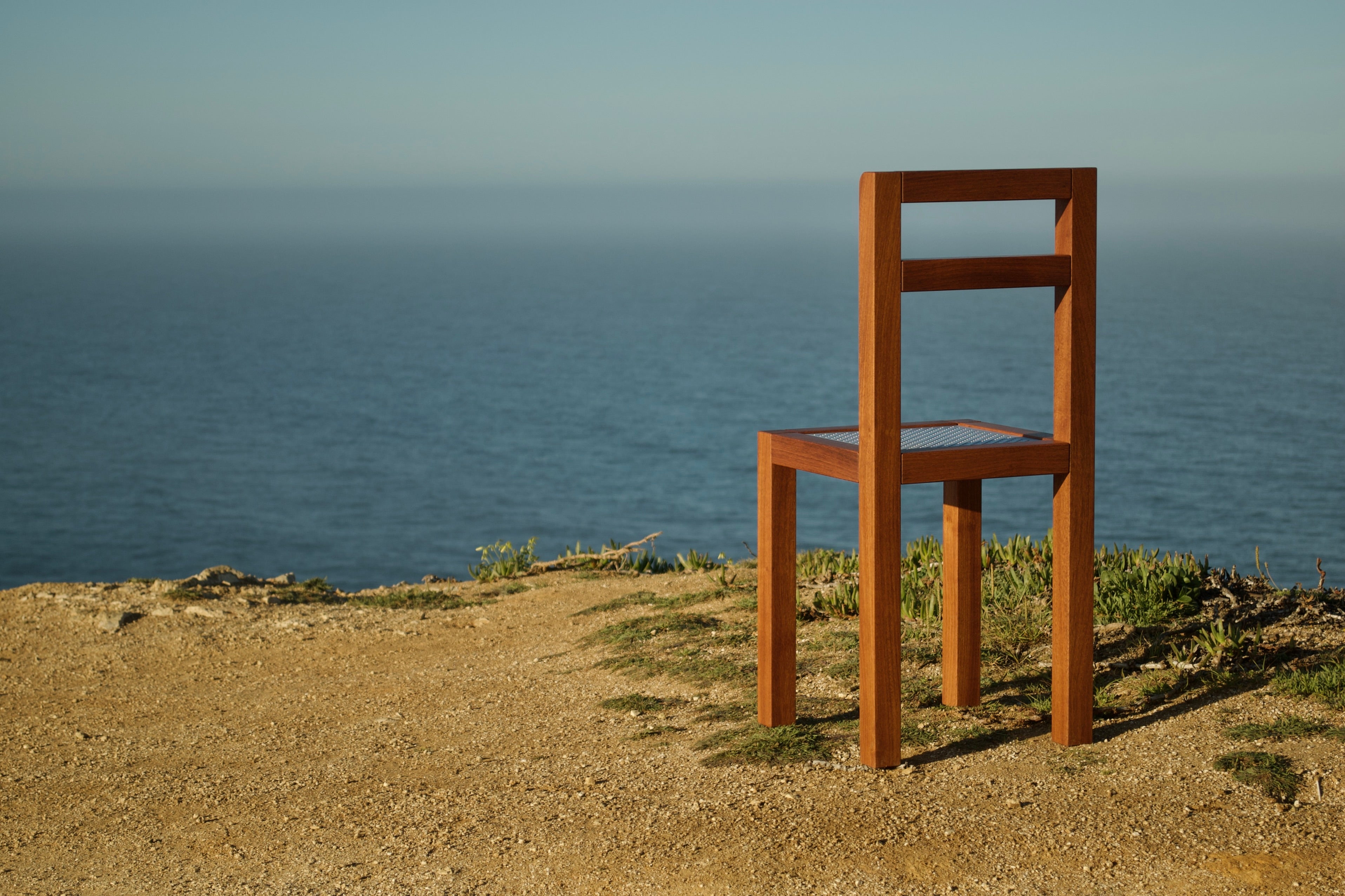 Modern minimalist designer wooden chair with a perforated metal seat. Back view with the ocean in the background.