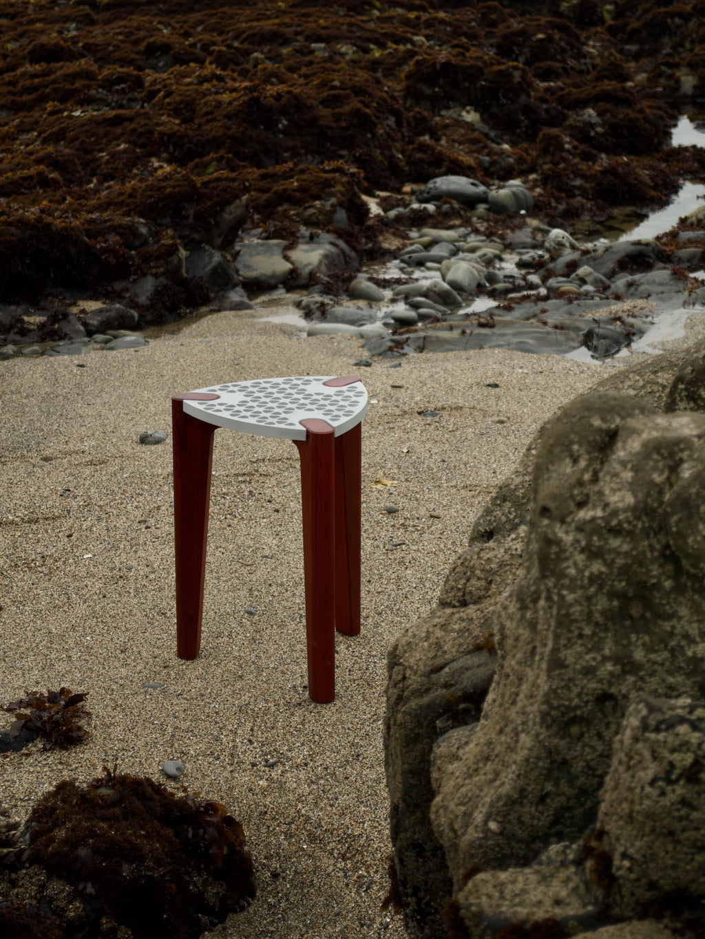 Modern minimalist designer stool/table with an aluminum top and red hardwood legs. Top-side view from a distance with an ocean beach in the background.