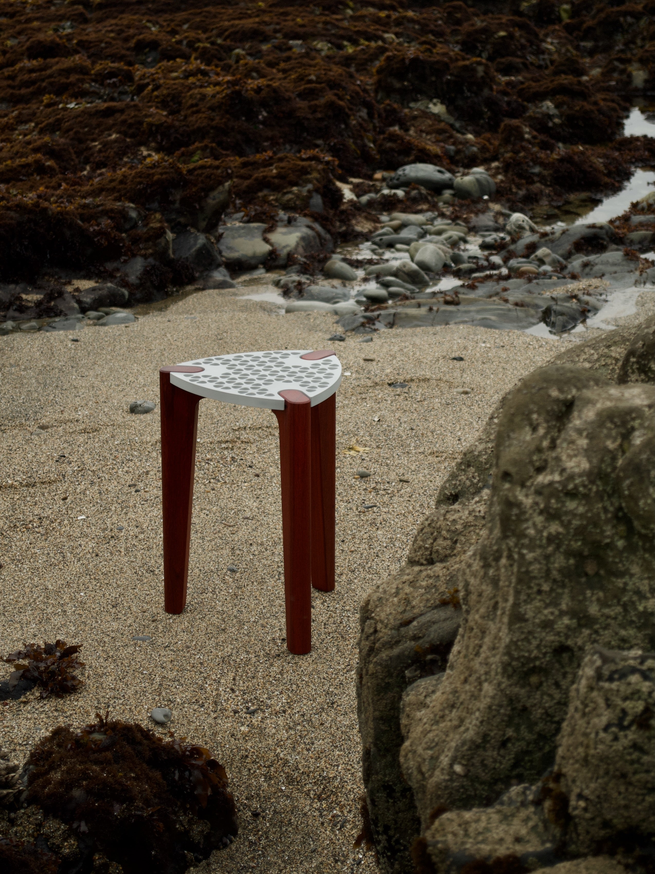 Modern minimalist designer stool/table with an aluminum top and red hardwood legs. Top-side view from a distance with an ocean beach in the background.
