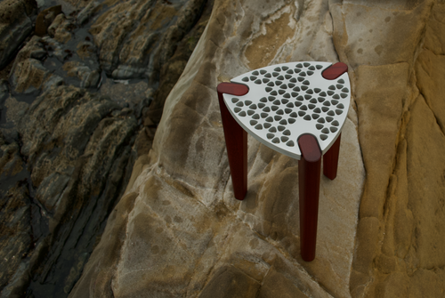 Modern minimalist designer stool/table with an aluminum top and red hardwood legs. Top-side view on a rocky background.