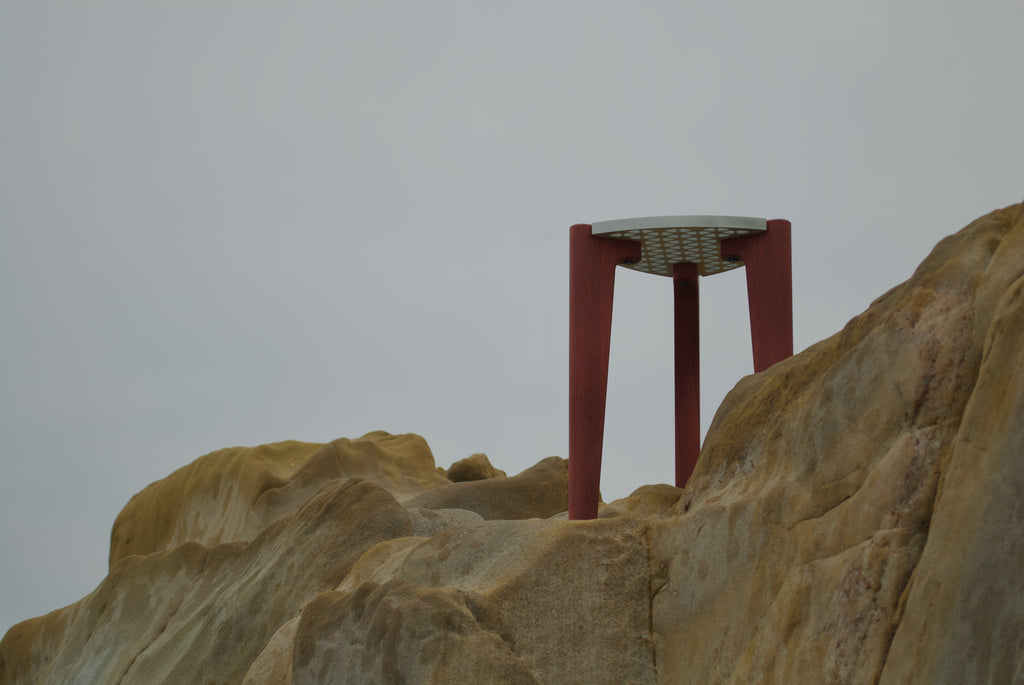 Modern minimalist designer stool/table with an aluminum top and red hardwood legs. Bottom-side view on rocks with the sky in the background.